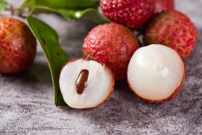 a bushel of lychee, a red, prickly skinned spherical fruit with white, glossy edible flesh surrounding a dark oval shaped seed. two of the lychees' skin is peeled to reveal the white flesh