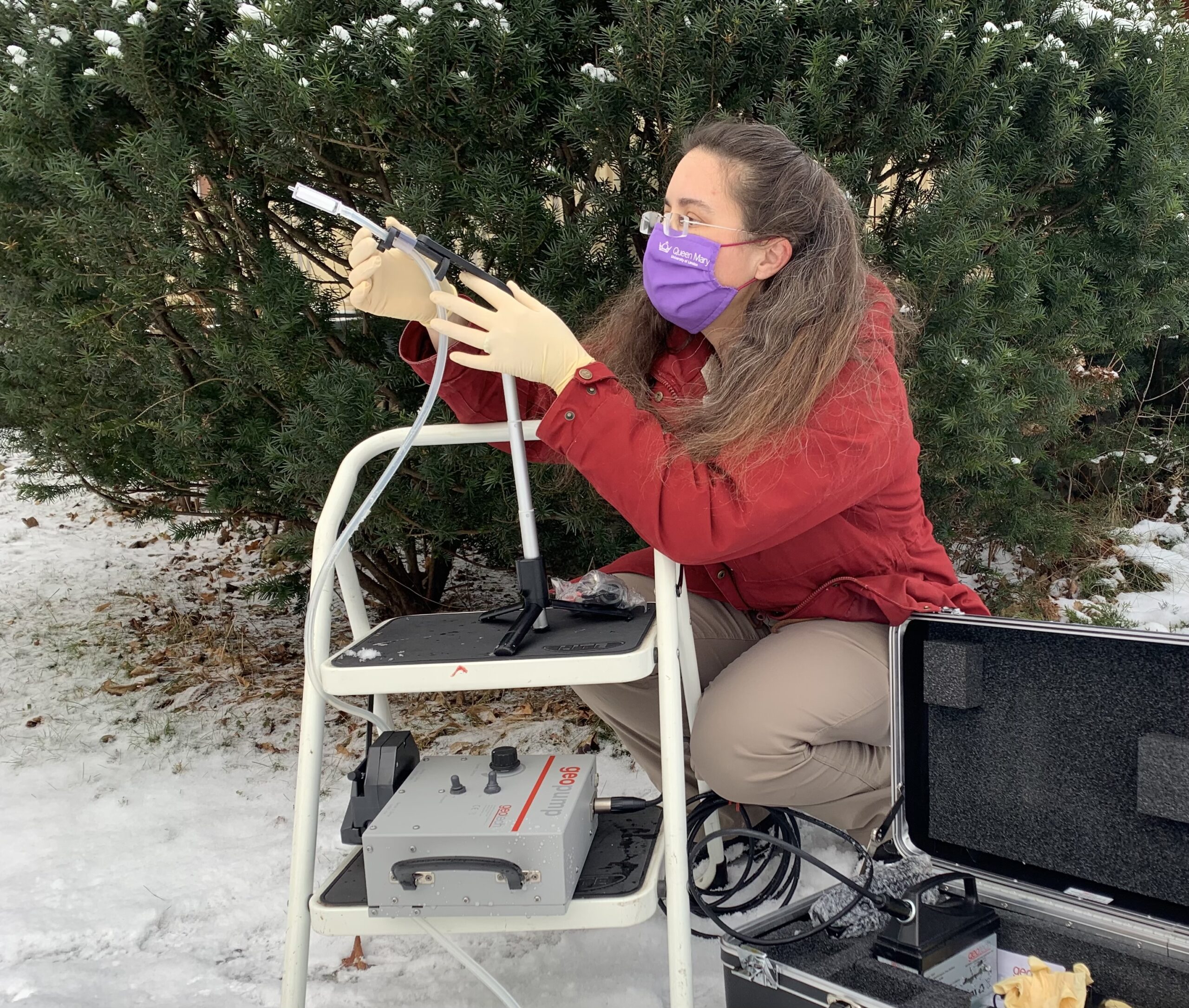 a woman in a purple face mask, gloves, and red jacket holds a piece of air collection equipment, which has a long tube feature. she is kneeling down next to a stool where the equipment is perched. behind her is a large green bush