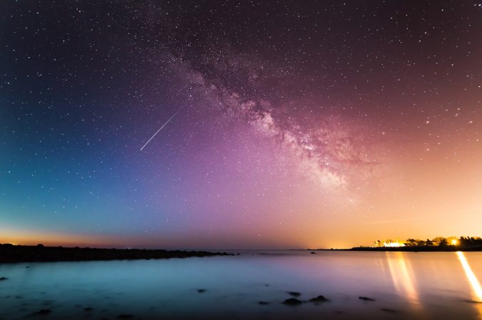 A shooting star in a purple, blue, and yellow night sky over water