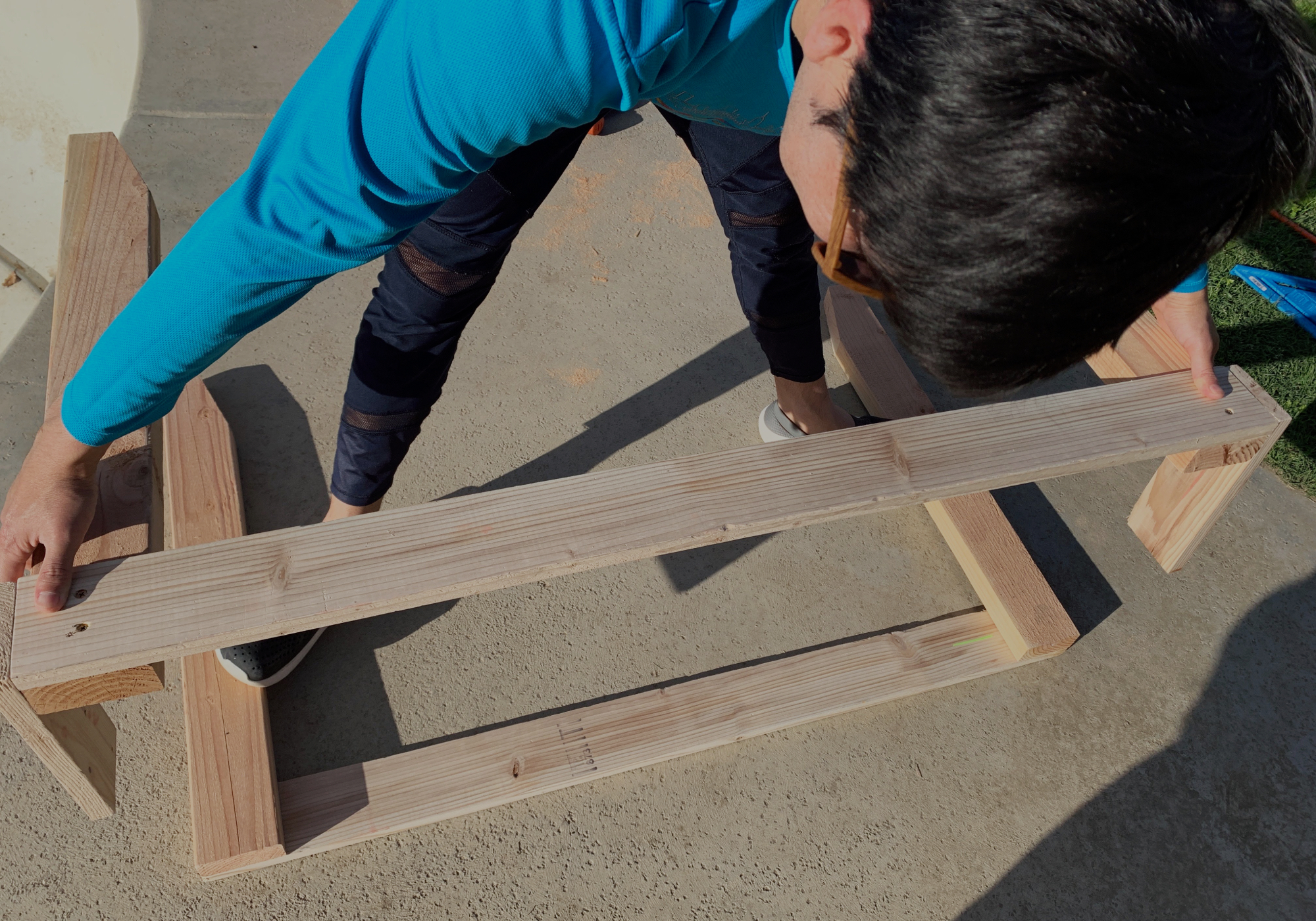 A person assembling a wooden DIY shoe rack outside on a concrete driveway.