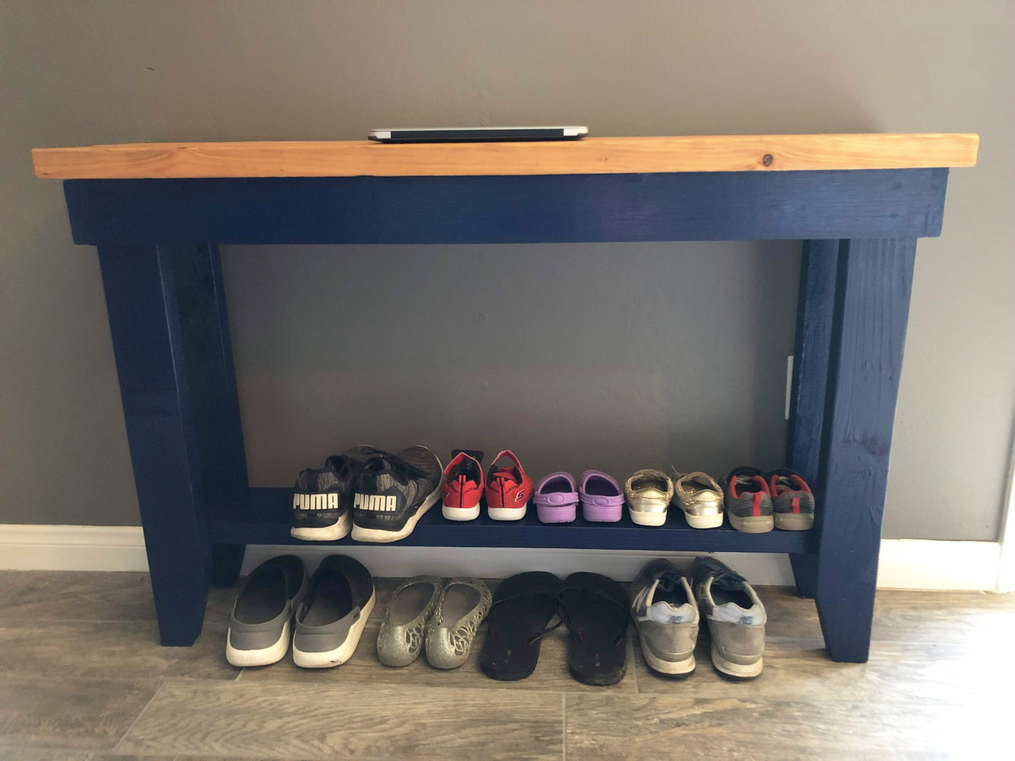 A blue wooden shoe rack table against a gray wall with neatly paired shoes on it.