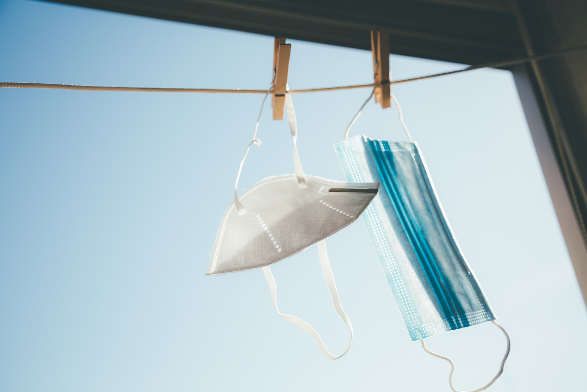 A white COVID respirator mask and blue surgical masks hanging on a clothesline in a window