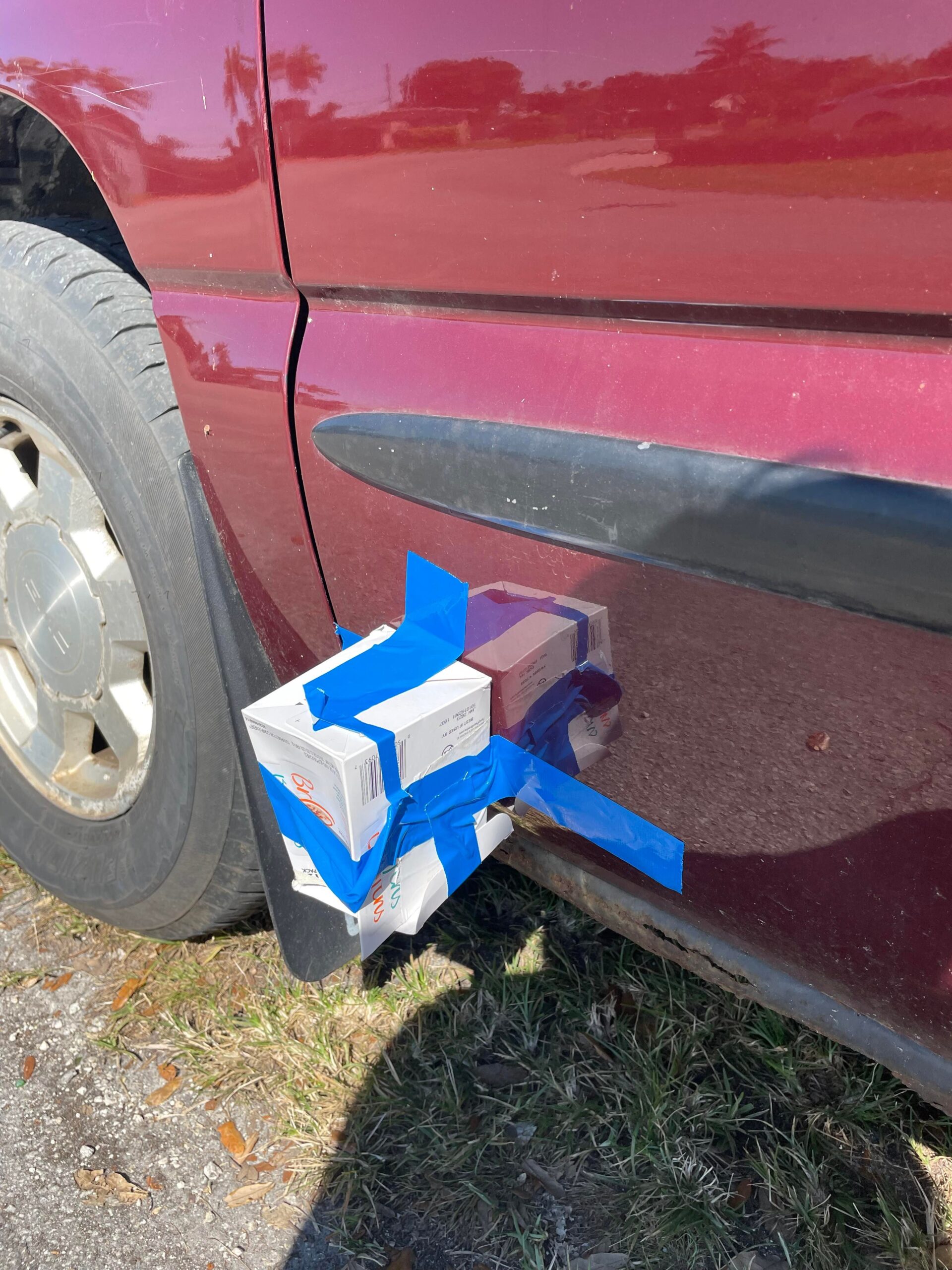 Carboard box with blue duck tape covering an umbrella plant on the driver's side door of a maroon GMC Sierra pickup truck