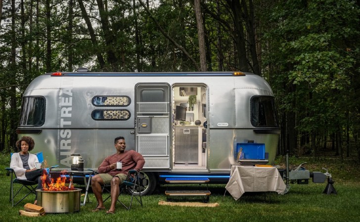 Black couple at a campfire in front of a silver Airstream RV trailer