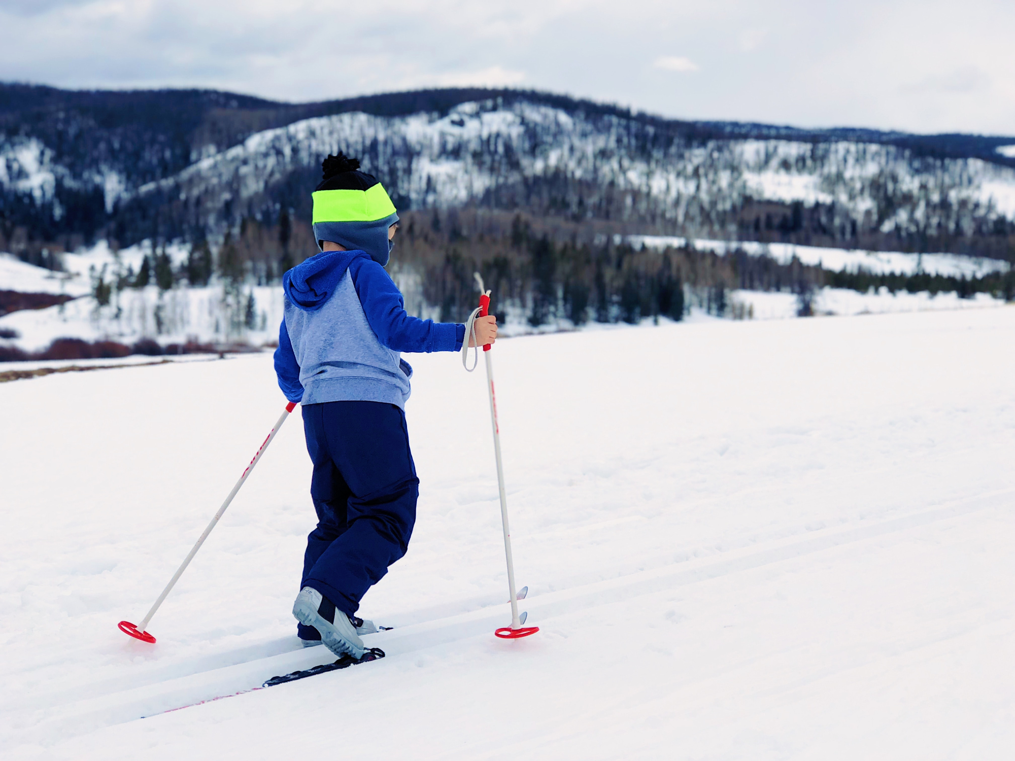 A child cross-country skiing on a snowy field near some round, tree-covered mountains.