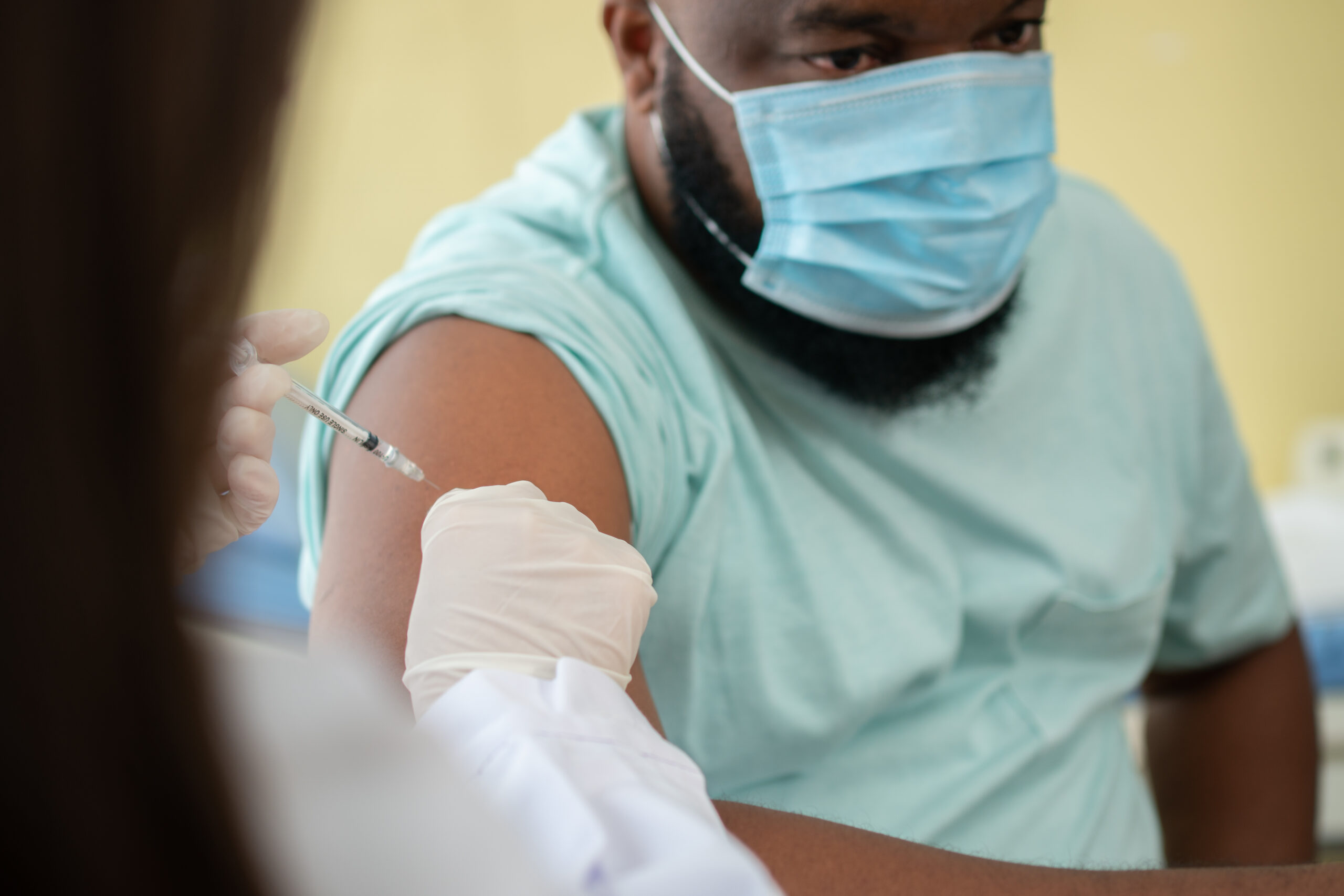 A man wearing a surgical mask is vaccinated by a healthcare worker wearing gloves.