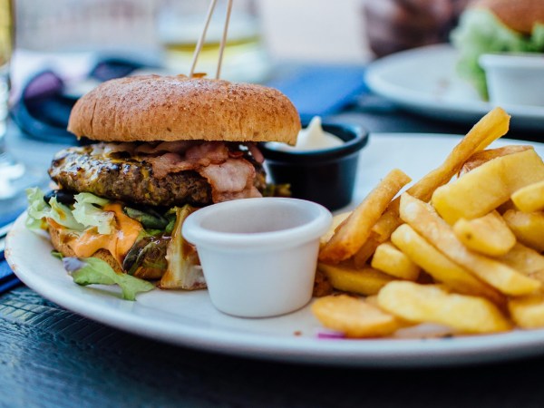 A burger with bacon, lettuce, and cheese on a white plate with two plastic containers of condiments and some French fries next to it.