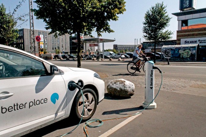 A white car sitting in a parking space being charged from a pole with a cable.