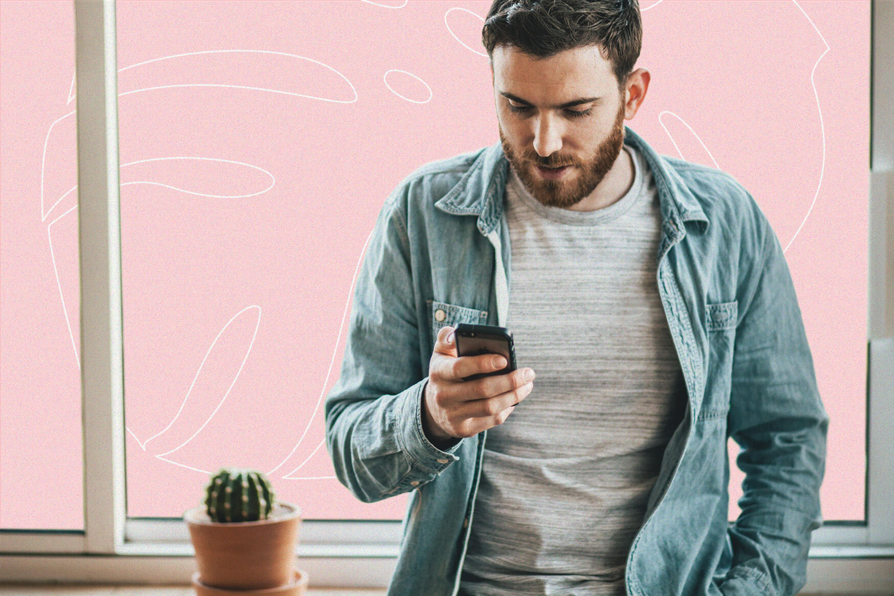 A man looking at his phone and leaning against a windowsill next to a potted cactus about the size of a softball.