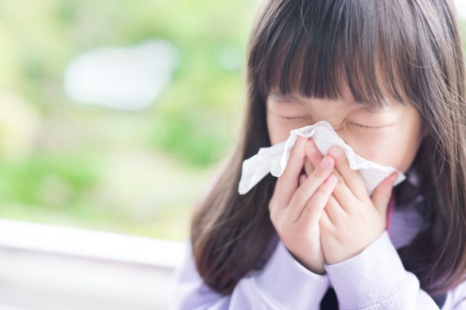 a little asian girl sneezes into a tissue with greenery in the background