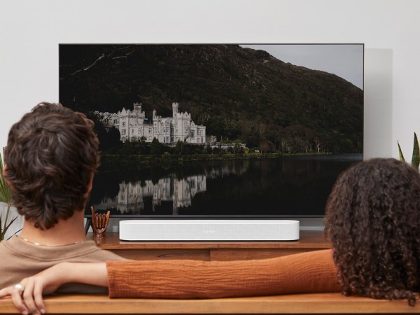 Two people, viewed from behind, sitting on a couch in front of a large flat-screen TV, with a white Sonos soundbar connected to it.