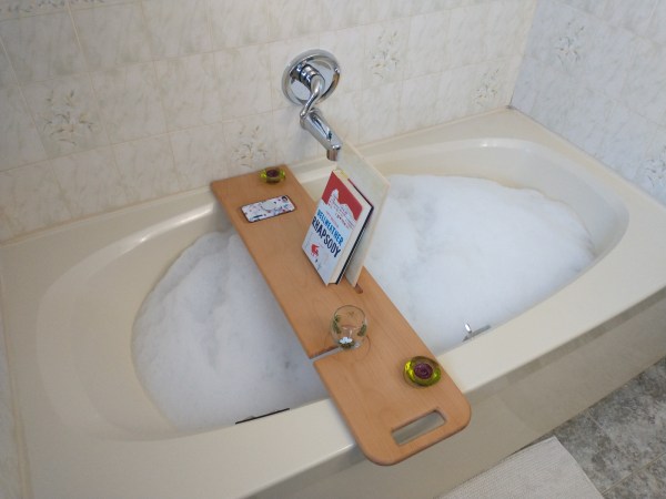 A bathtub full of bubbles with a homemade wooden bath tray on it, complete with a book, a phone, a candle holder, and a wine glass holder.