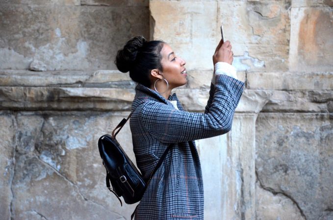 A woman standing in front of a stone wall and looking through her phone at something off-screen, maybe scanning text on a sign.