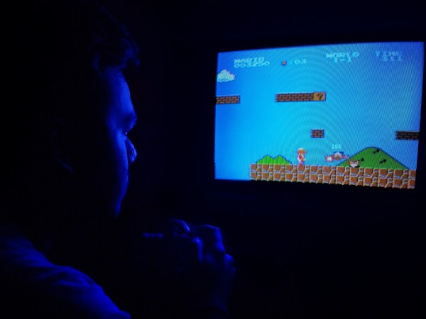 A man sitting in a dark room, his face illuminated only by the blue glow of a cathode-ray TV as he plays a retro Nintendo game.