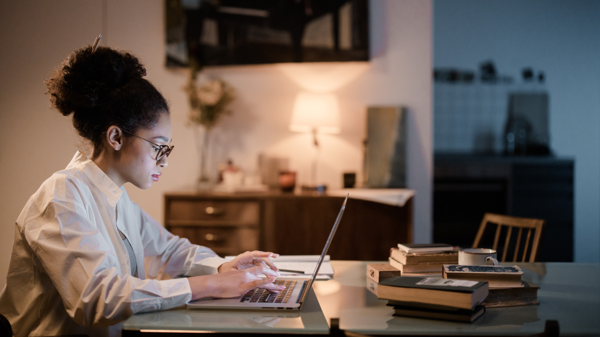 A young woman sitting at a table in a dimly lit room, typing on a computer with a lot of books on the table.