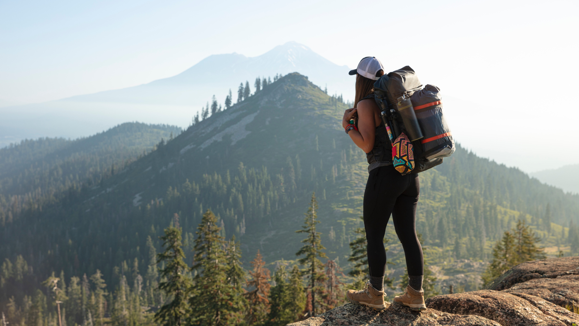 A woman in hiking attire and a large backpack standing on a cliff in a mountain range.