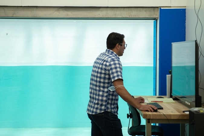 Ocean simulator in giant saltwater tank at the Scripps Institute in San Diego, California. A Scripps researcher in a checkered shirt adjusts the settings on a computer with a COVID mask around his elbow.