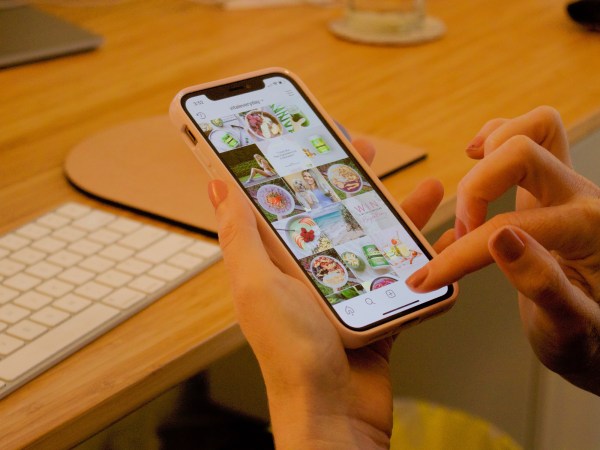 A person sitting at a light wooden desk in front of an Apple desktop computer, scrolling through Instagram on their iPhone.