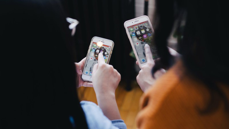 Two black-haired women, from the back, using their iPhones with each other.