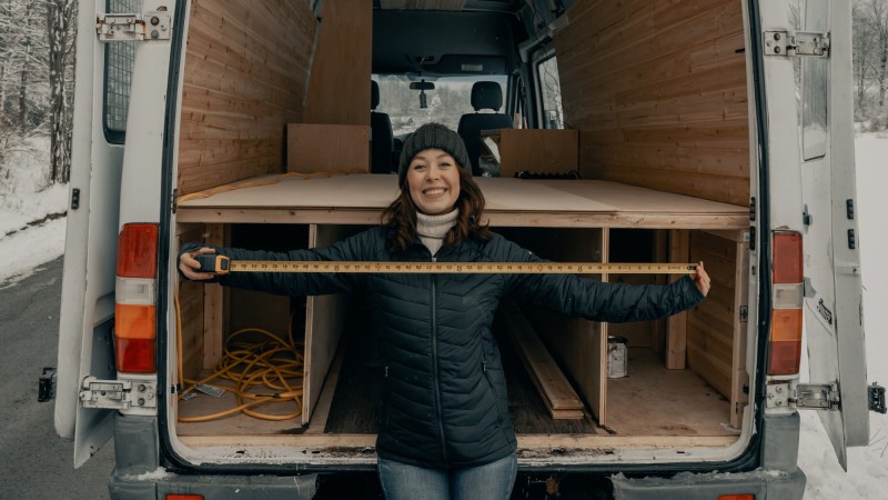 A woman standing at the back of a partially completed van conversion project, holding her arms out wide with a measuring tape held between them.