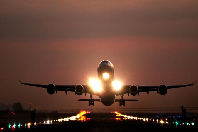 An airplane taking off toward the camera at dusk, with lights along the runway and on the front of the plane, against a cloudy reddish sunset.