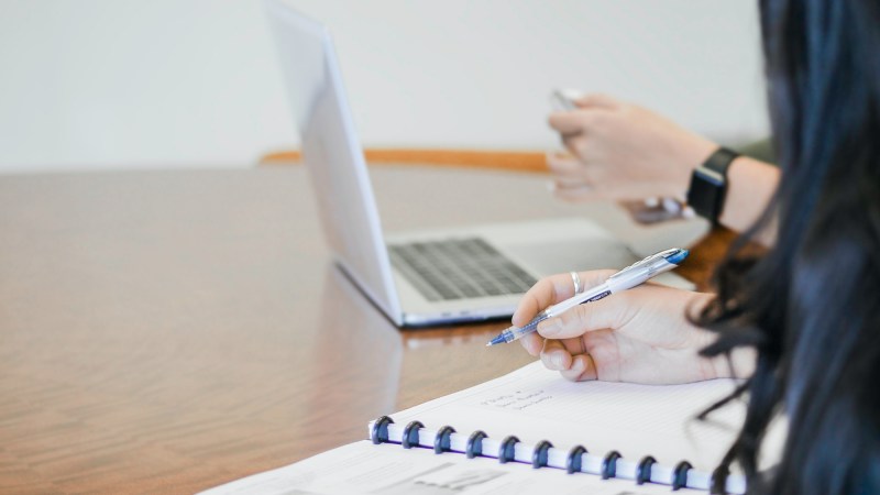 Two people sitting at a wooden table in an office space, with one of them writing in a paper calendar, and the other looking at a phone while working on a laptop.