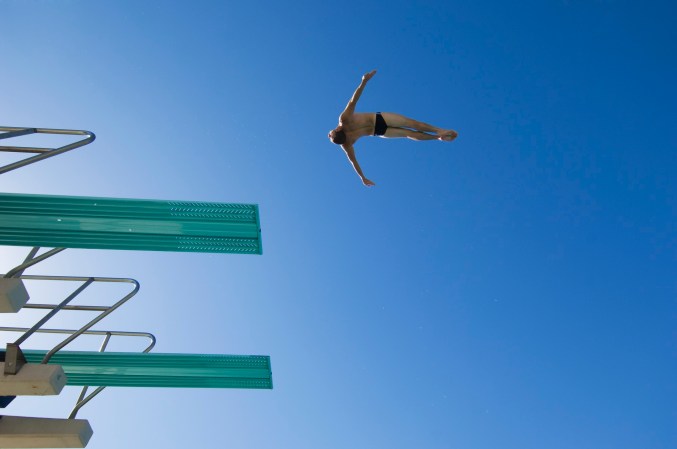 Swimmer in a black speedo soaring off a green diving board