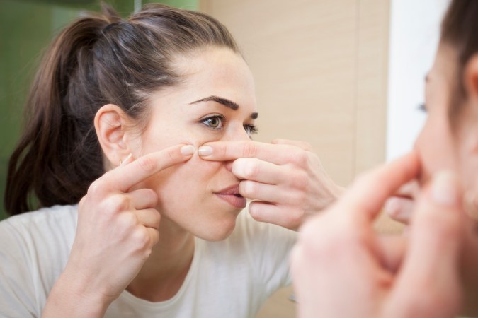 a woman looks in the mirror as she pinches a pimple on her check with her two pointer fingers