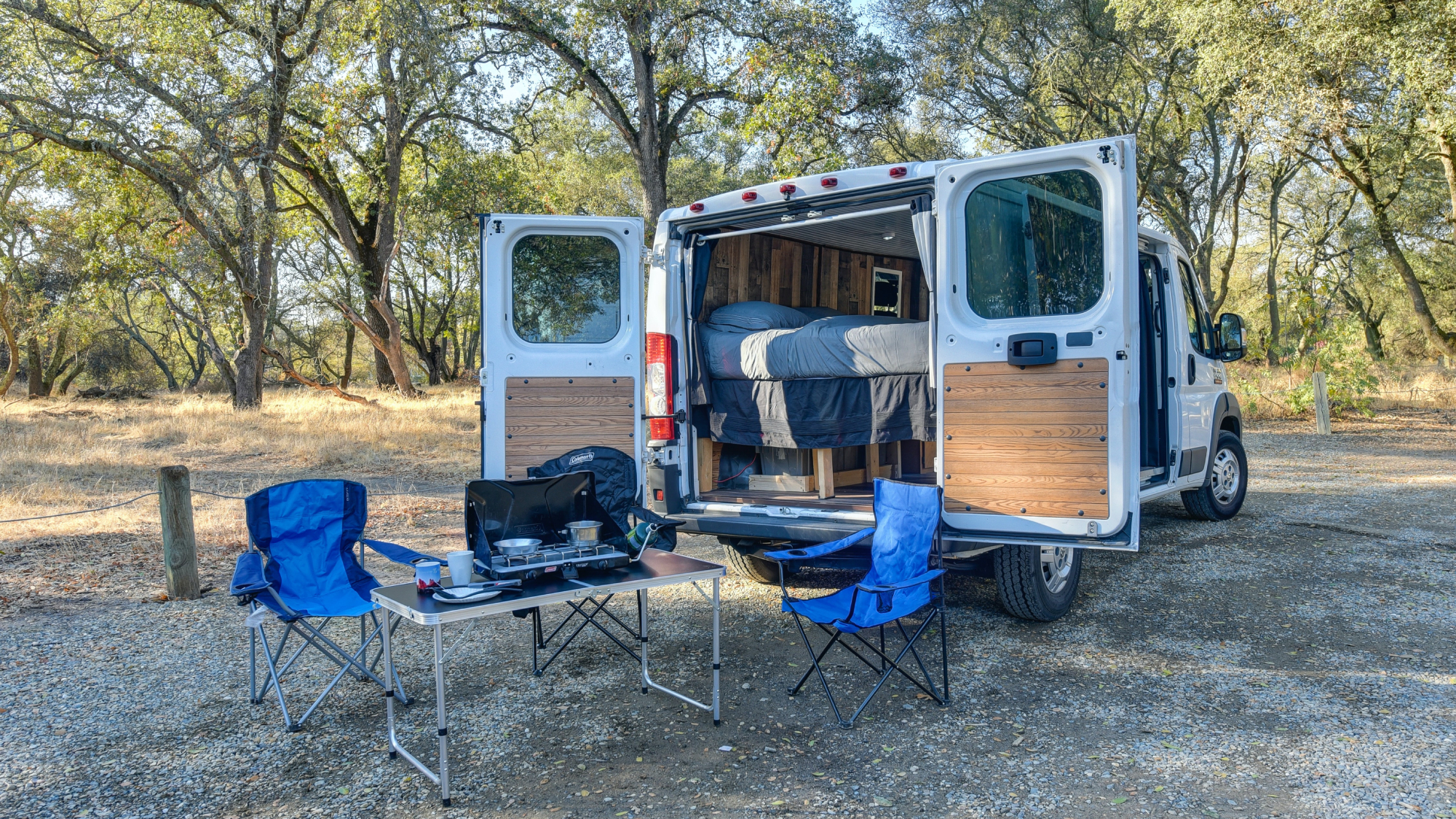 A van-life van with its back doors open and two camping chairs and a small grill set up behind it.