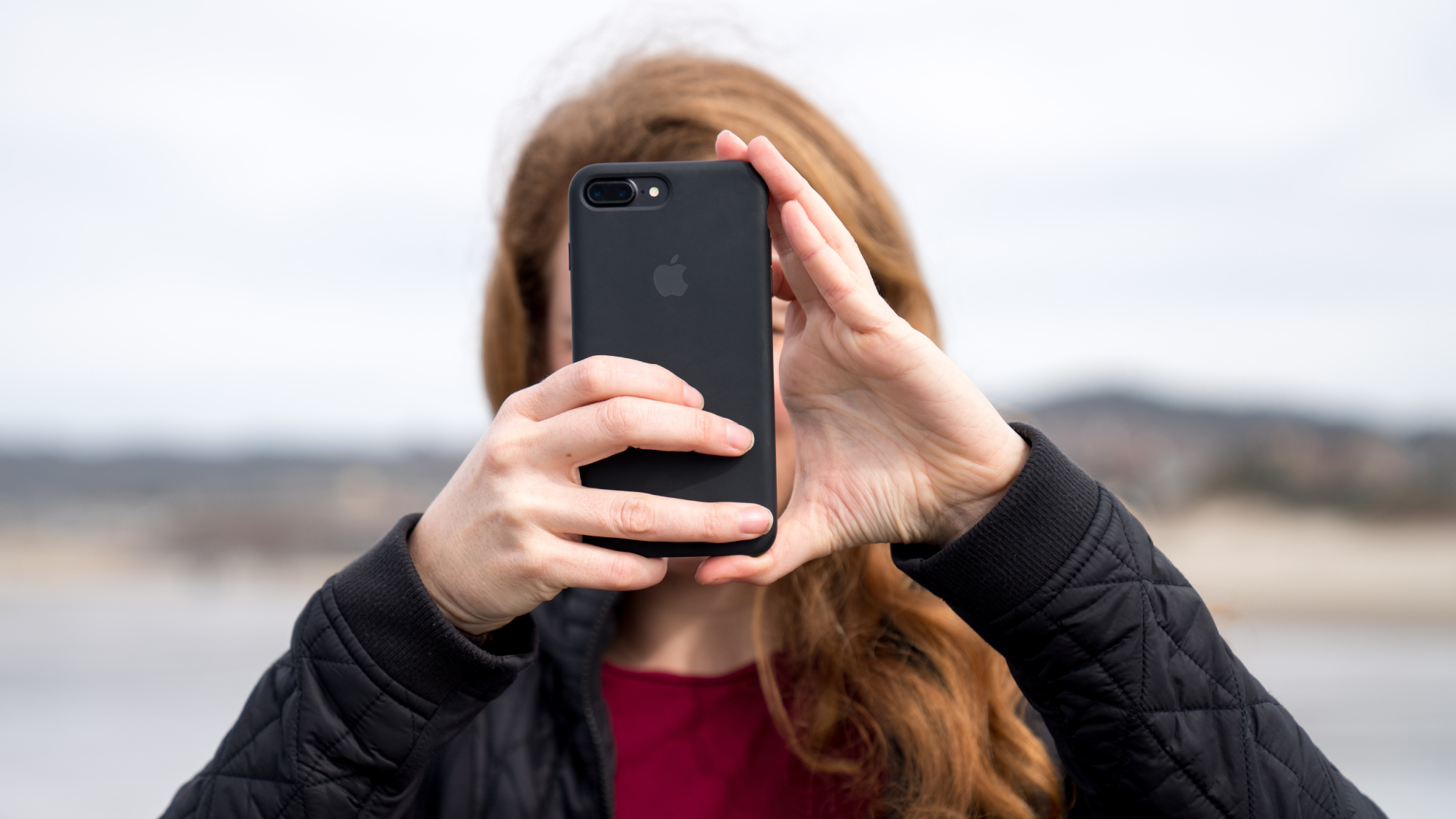 A red-haired woman facing the camera, using her black iPhone to take a photo, with the phone covering her face.