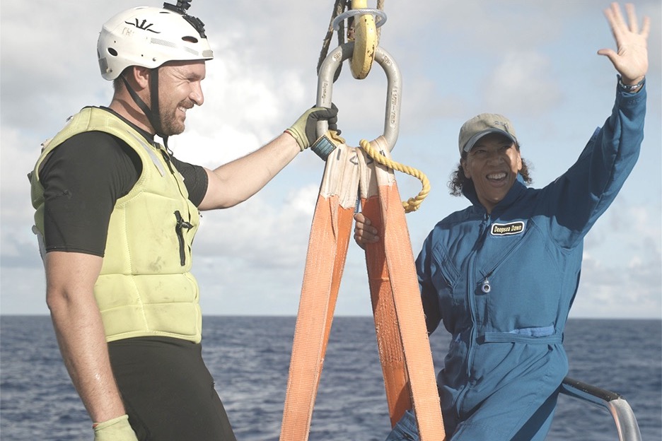 a black woman wearing diving gear and a hat waves excitedly next to a man holding ropes to the sub steady, as she lowers into the sub