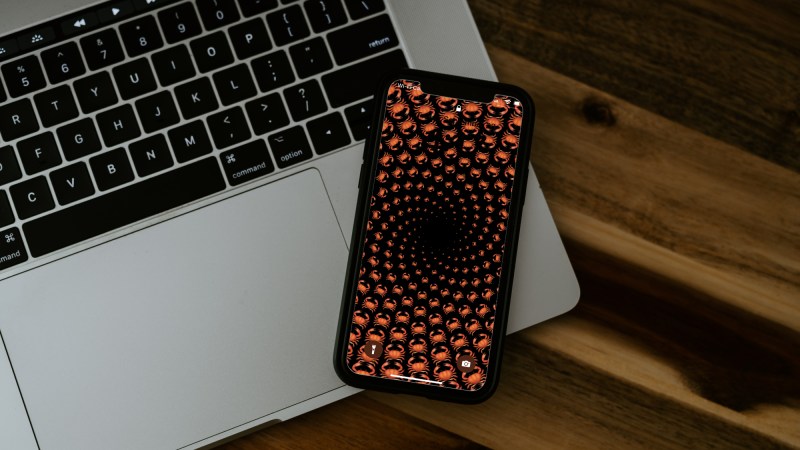 An iPhone on top of a Macbook laptop on a wooden table, with the iPhone displaying a spiral of crab emojis, which is one way you can edit your iOS 16 lock screen, if you're into that.