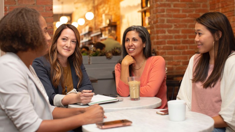 Four women sitting around two small, white, round tables having a conversation.