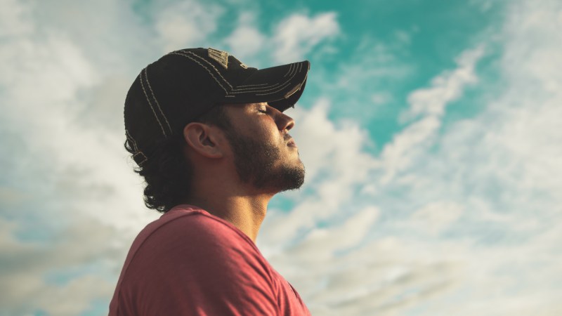 A man standing under a blue sky with white clouds, wearing a black baseball cap and a red shirt while closing his eyes and breathing deeply.