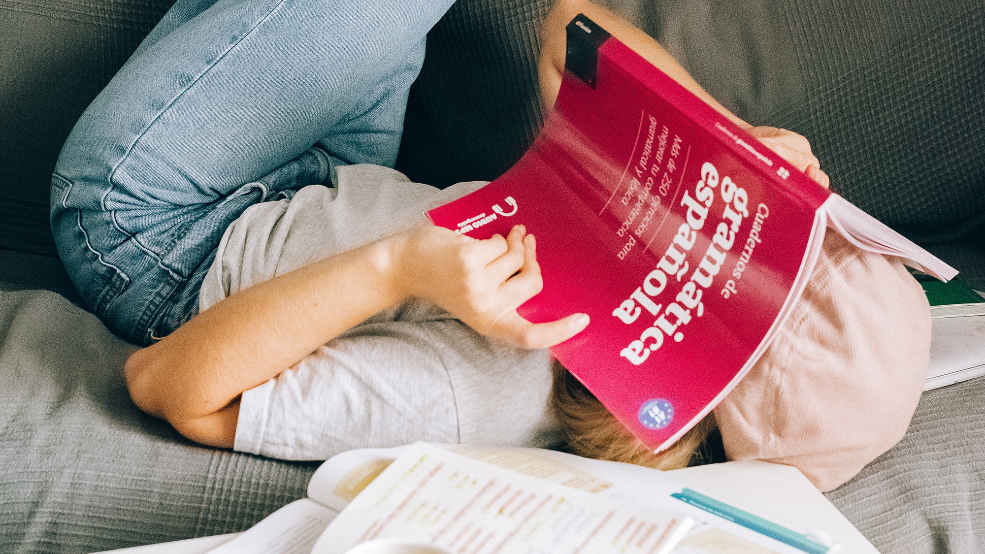 Person lying on couch covering their face with a book about Spanish grammar