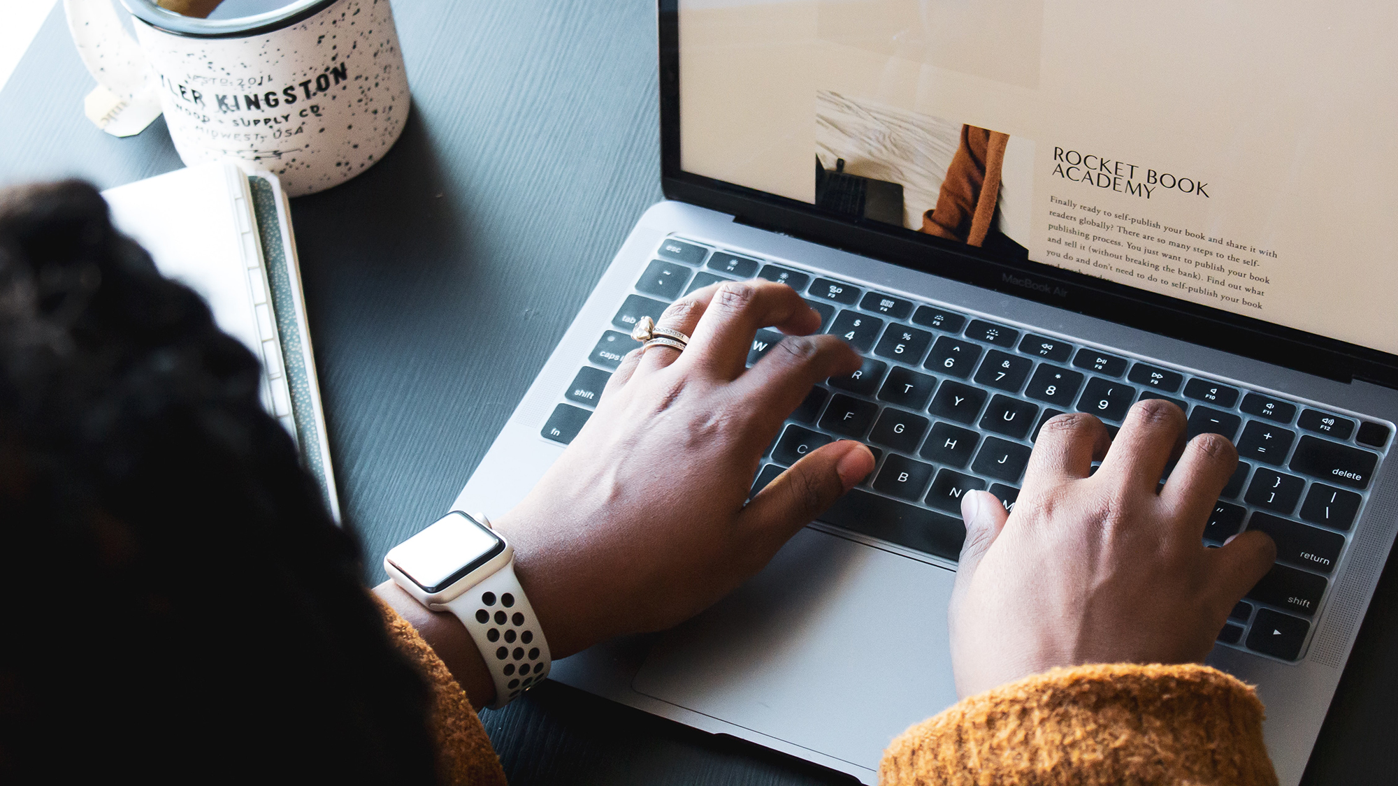 hands on laptop typing next to a notebook and a coffee mug