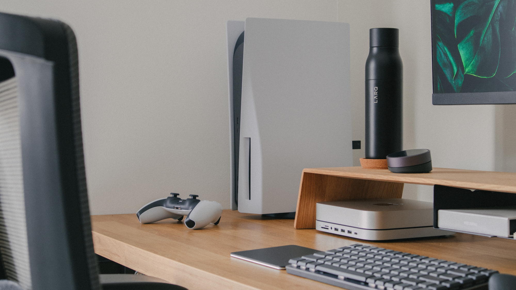 A PS5 on a wooden desk with a DualSense controller, a keyboard, and a mouse nearby.