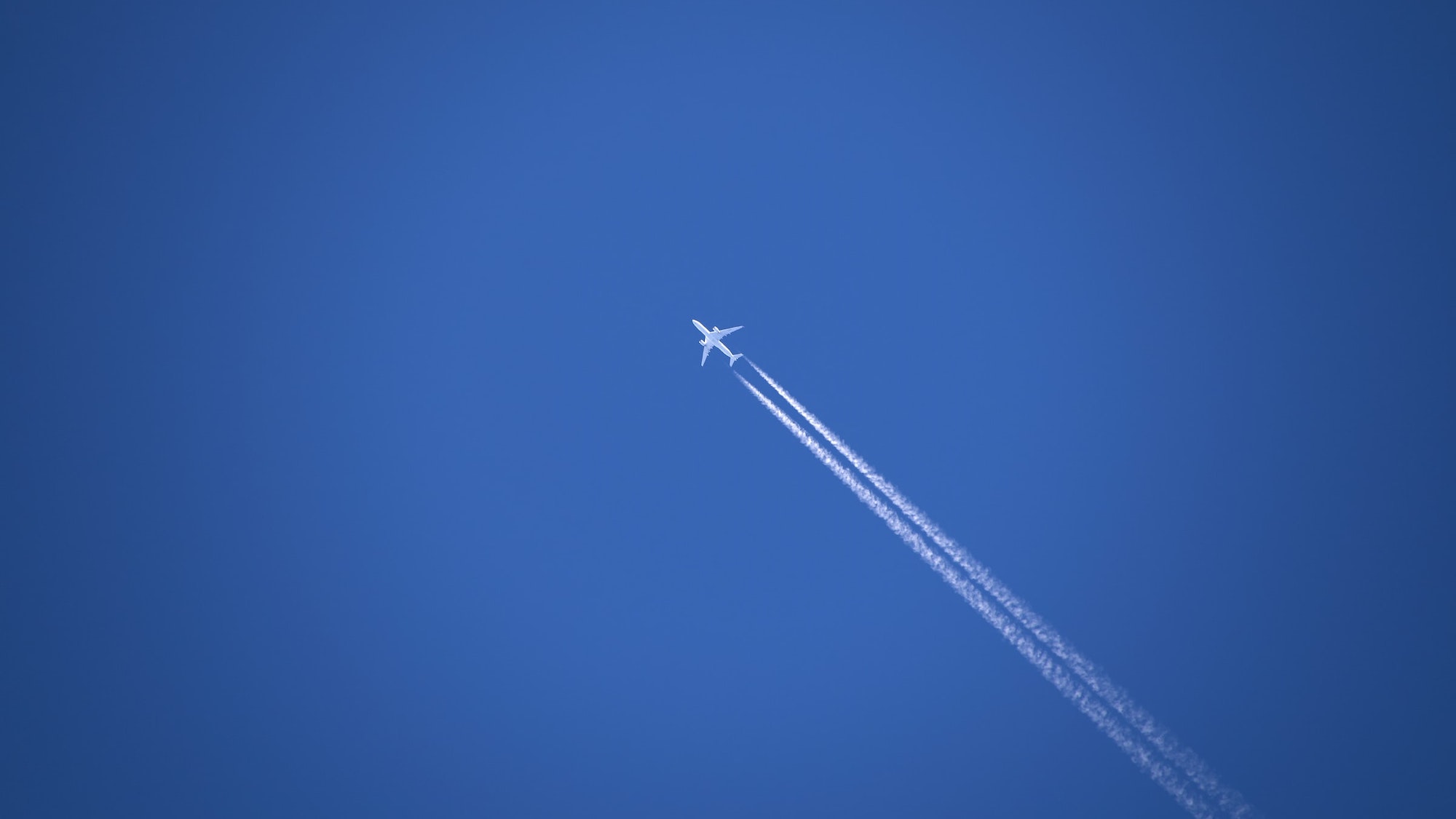 A plane flying high above the ground, drawing white contrails across a blue sky.