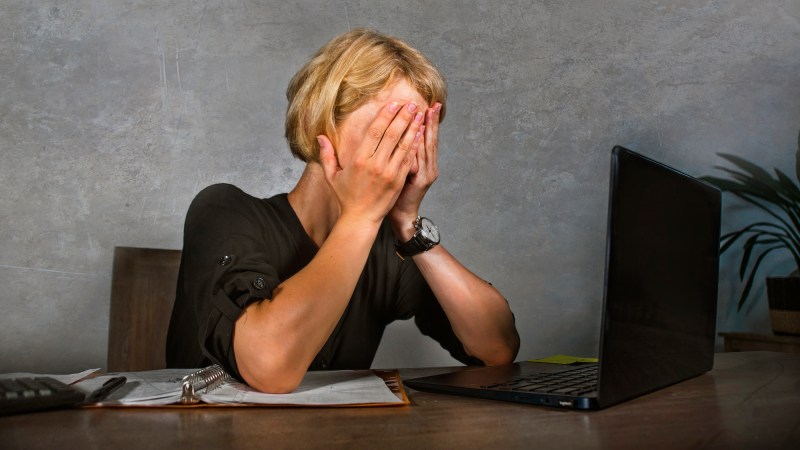 Woman covering face with hands in frustration while sitting in front of laptop on an office desk.