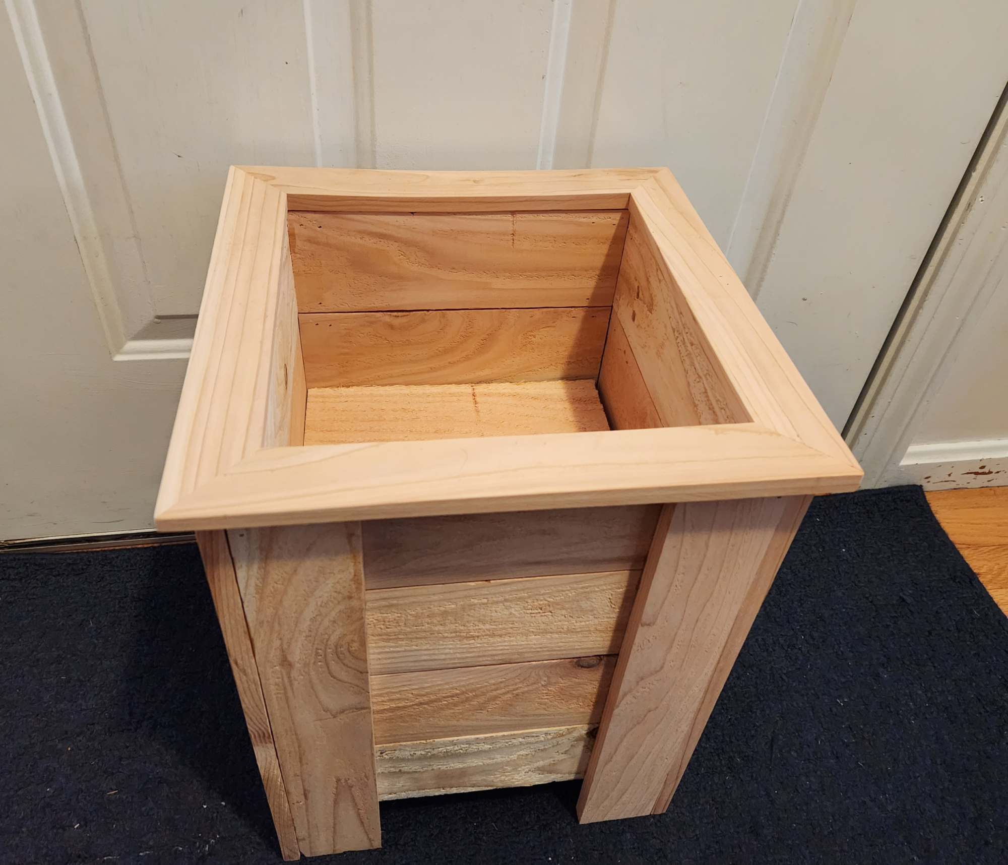 An empty cedar DIY planter box on a blue carpet in front of a white door inside a house.