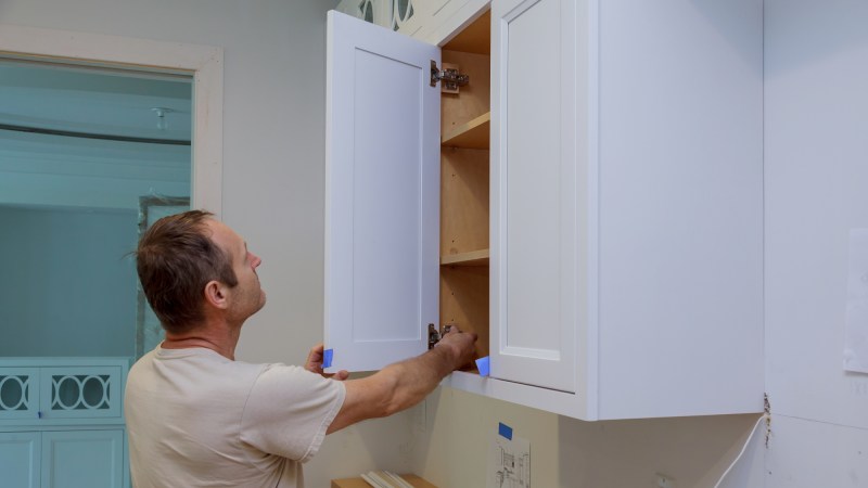 A man with short, brown hair, wearing a white t-shirt and hanging a white cabinet door in a white kitchen.