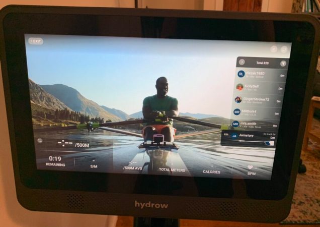  The touchscreen of the Hydrow Wave Rower, which features a world-class rower on a river with mountains in the background demonstrating how to row.