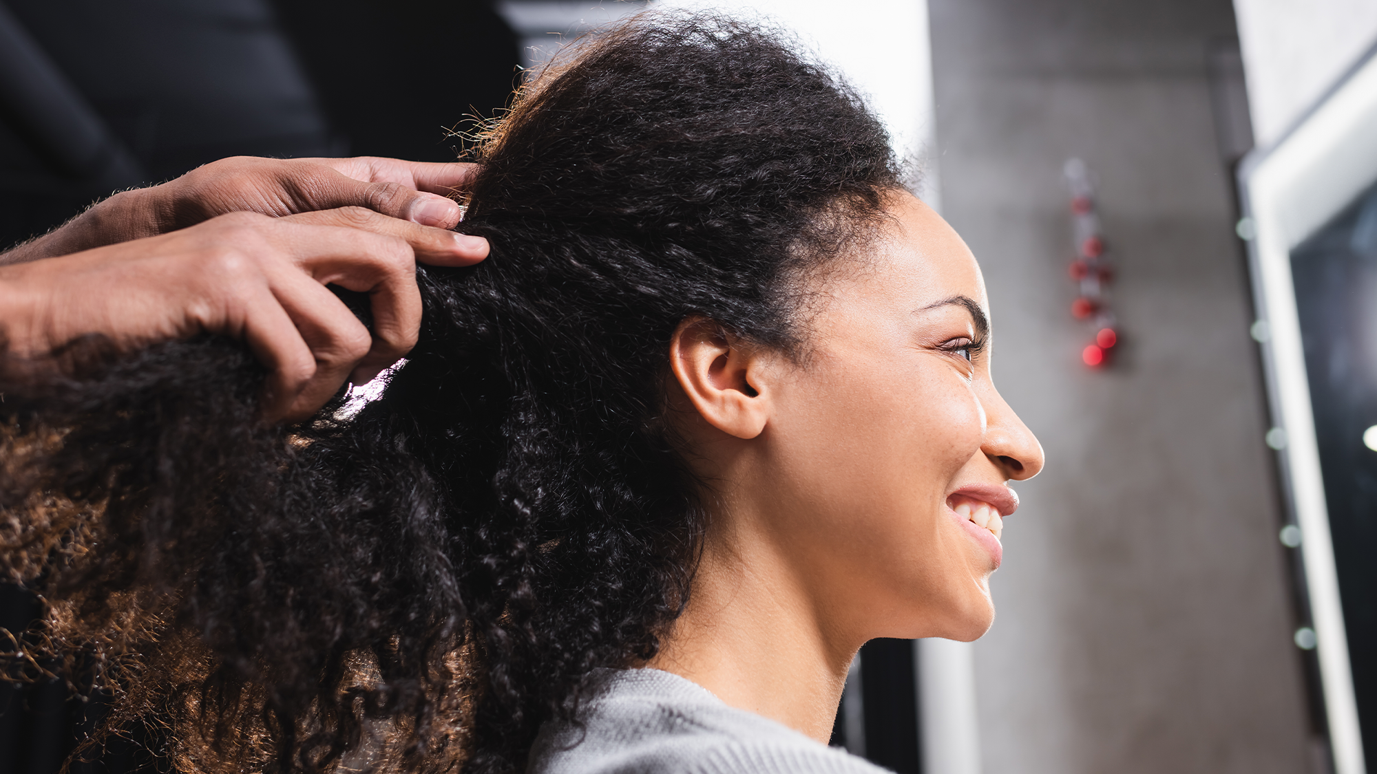 A Black woman smiles into a mirror at a salon for hair care.