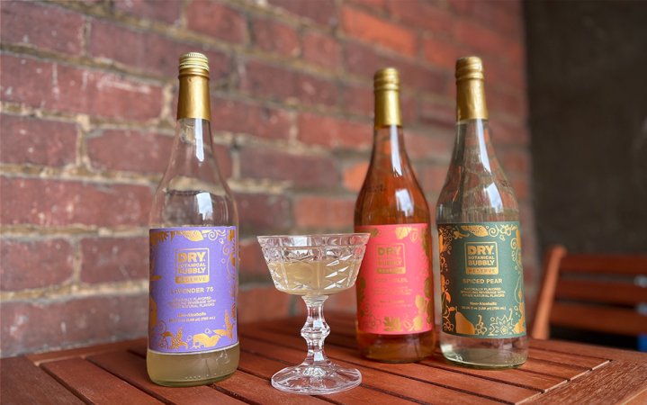  A lineup of DRY Botanical Bubbly Reserve bottles on a wooden table in front of a blue background.