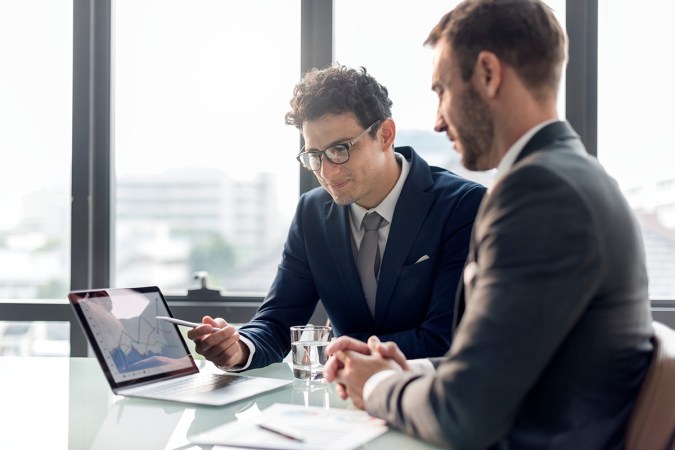 Two people wearing suits sitting at a desk talking