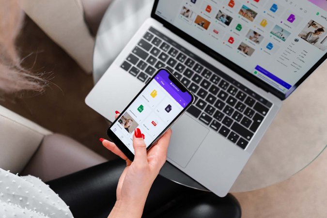 A person sits at a desk while holding their phone in front of their computer