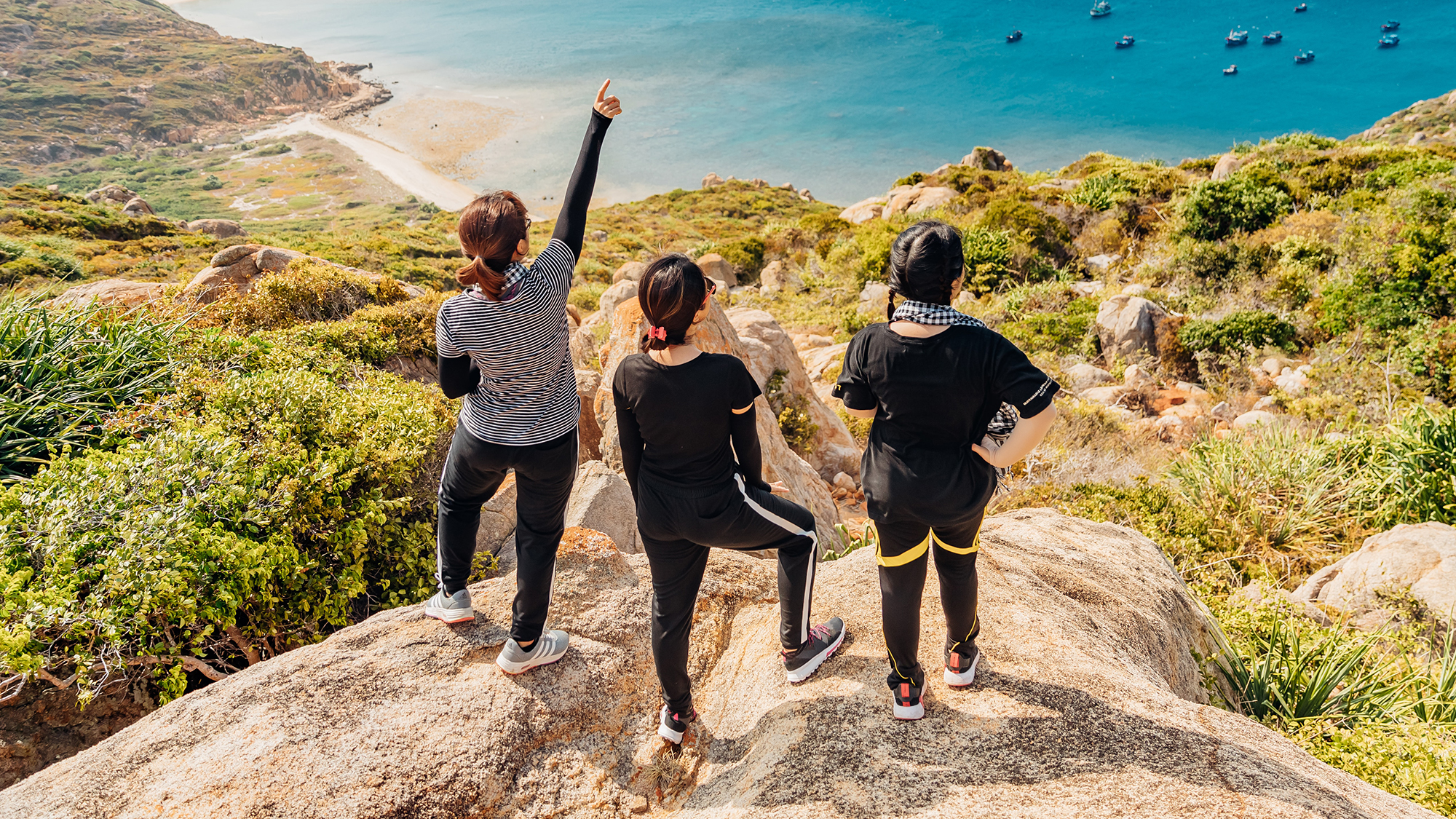 Three people standing on a high rock, looking at a beach downhill.