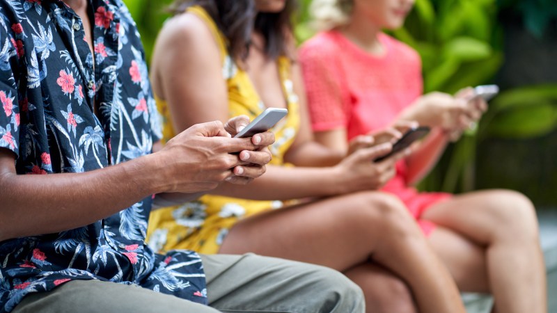 Close up of one one man and two women using their phones on a bench.