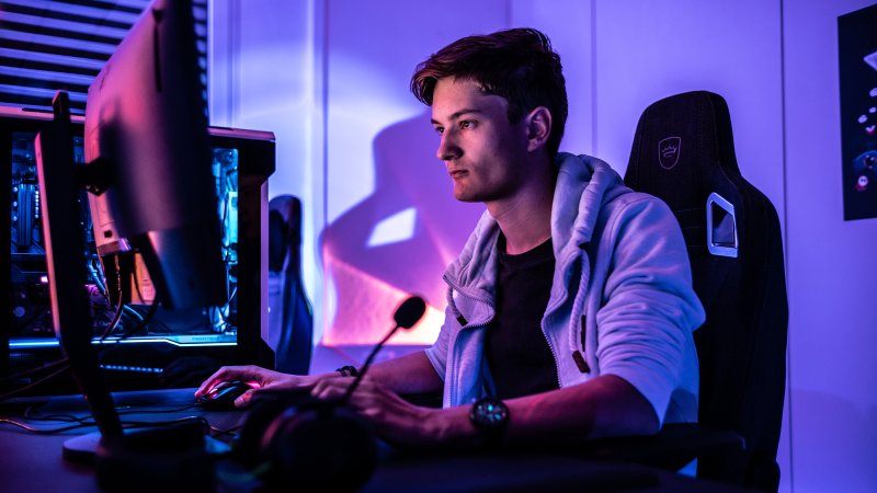 A person sitting in a dark room playing video games with a two-monitor computer set up.