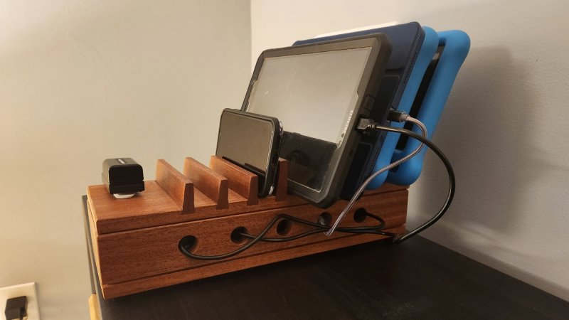 A wooden DIY device charging stand holding three tablets and an iPhone, on a black shelf against a white wall.